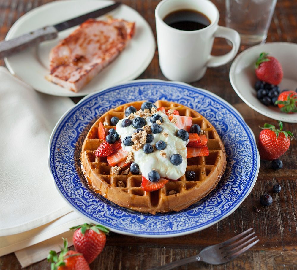 Round waffles with whipped cream, strawberries and blueberries next to a cup of coffee. Original public domain image from…