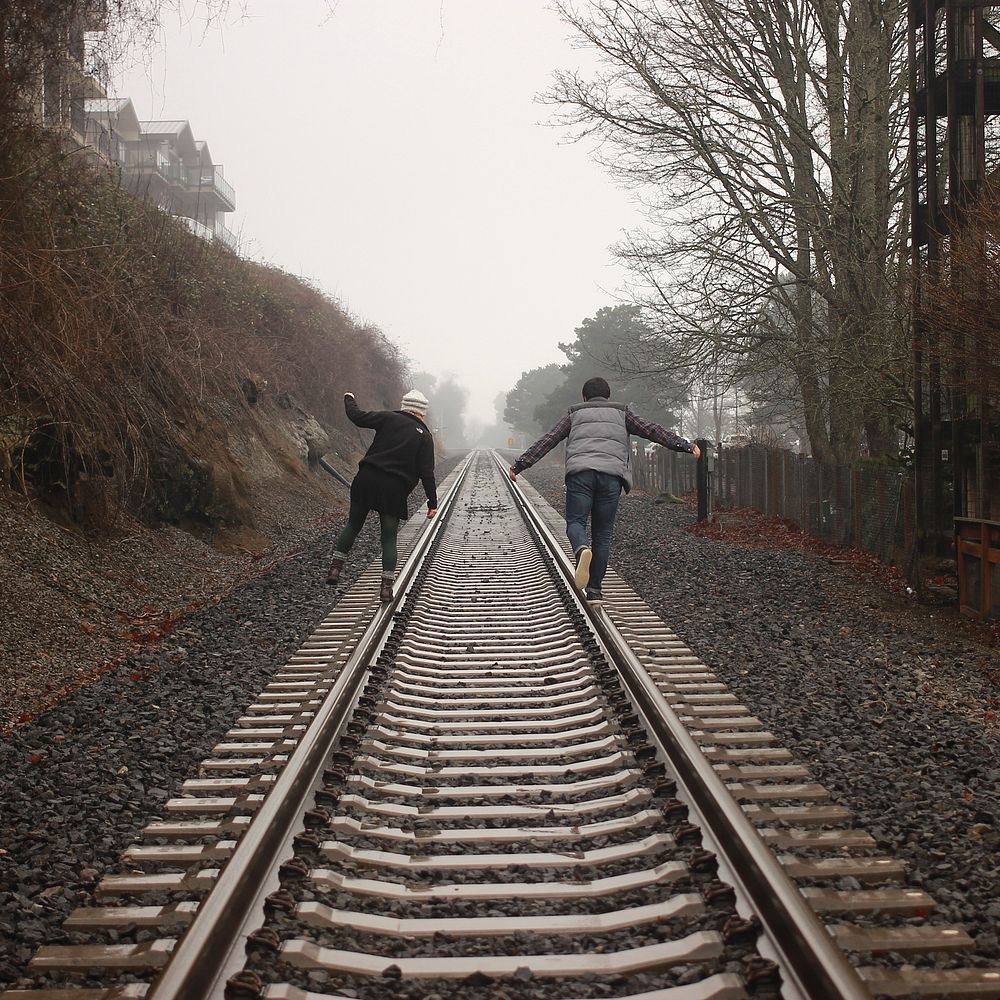 Two people balancing on the rails of a train track during a foggy day. Original public domain image from Wikimedia Commons