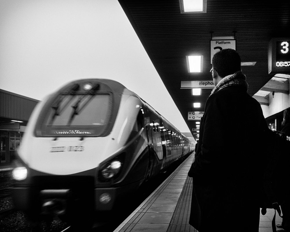 Black and white behind shot of man in coat with scarf standing on Leicester train platform. Original public domain image…
